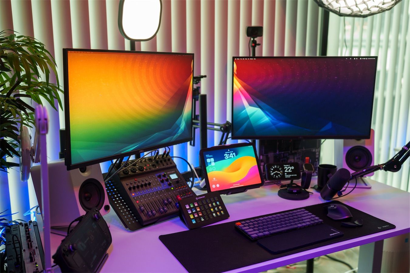 My white standing desk with two monitors (with colorful backgrounds), 2 white speakers, a mixer, an ipad on a stand and Elgato lights